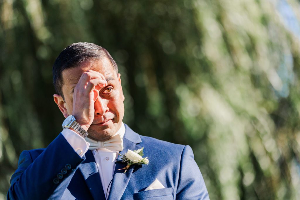 Groom wiping away tears during an emotional moment at the wedding ceremony, wearing a navy suit and white bow tie with a blurred greenery background.