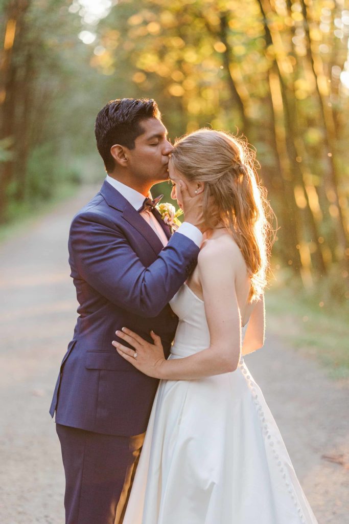 Groom in blue suit, kissing bride, and white dress on the forehead