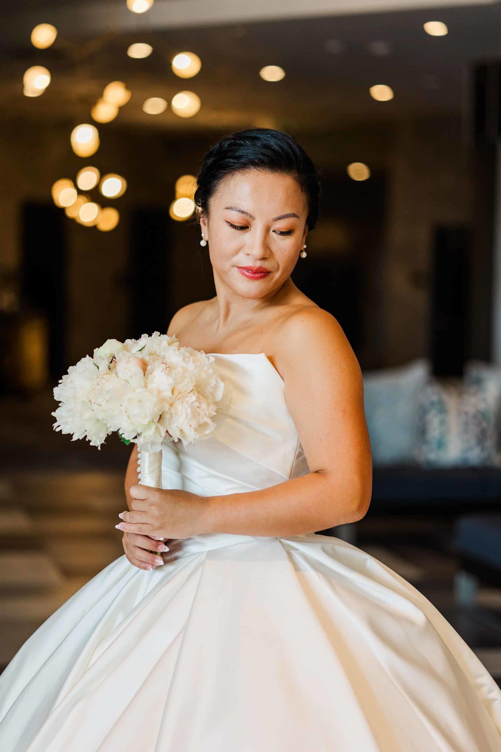 Asian, American woman holding a wedding bouquet looking down at the floor wearing a white dress