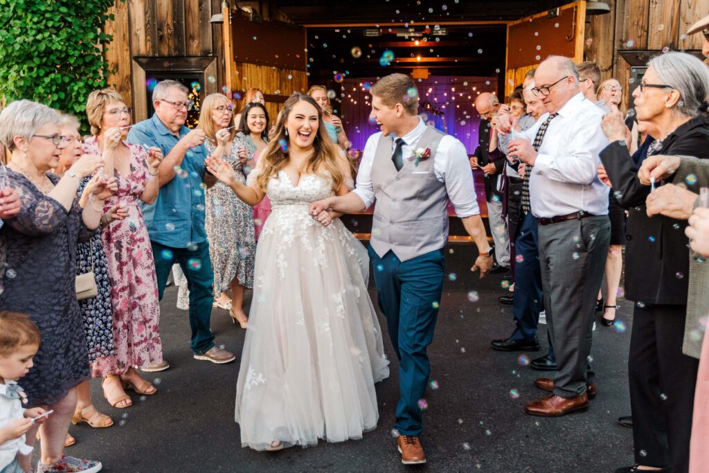 Bride and groom joyfully walking through a bubble-filled send-off surrounded by cheering guests outside a rustic wedding venue.