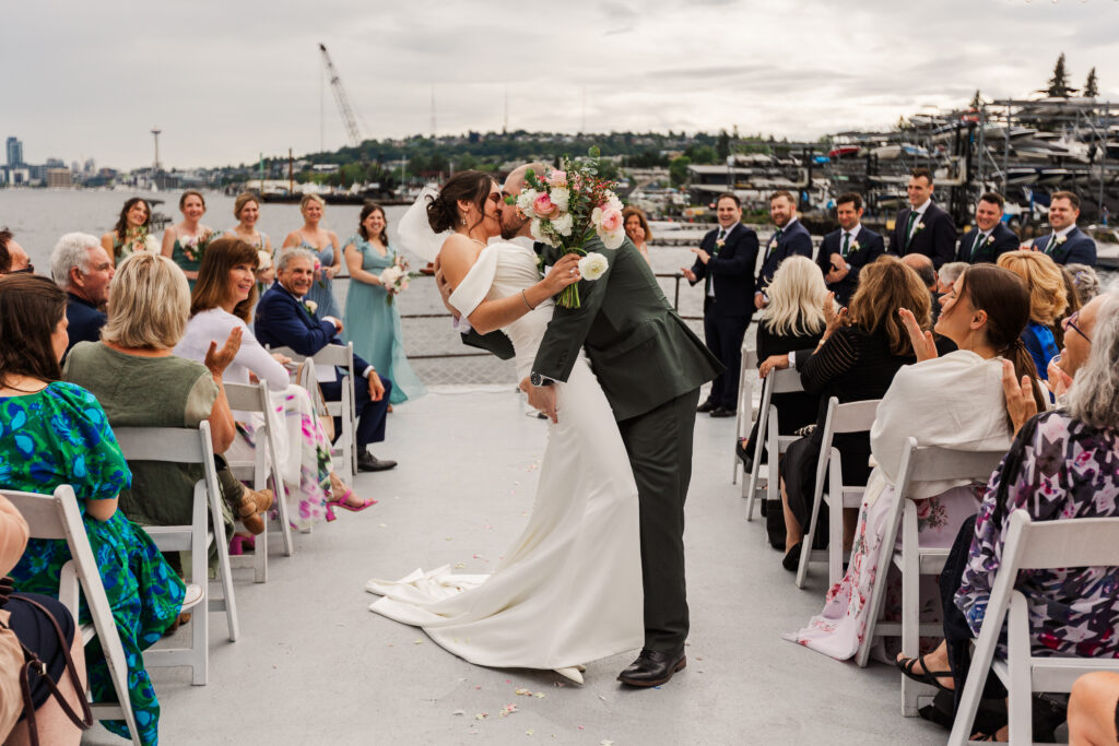 Bride and groom share a romantic kiss at an outdoor waterfront wedding ceremony in Seattle, surrounded by cheering guests and the iconic Space Needle in the background.