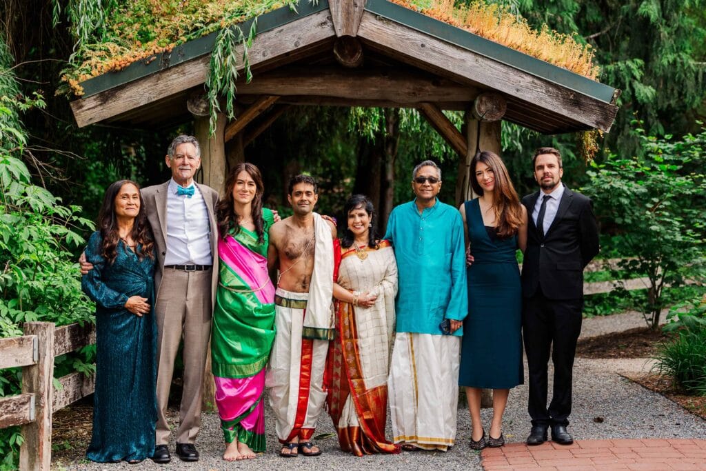 Family group portrait at an outdoor wedding, featuring members dressed in a mix of traditional South Asian attire and formal Western clothing, posed under a rustic wooden pavilion surrounded by lush greenery.