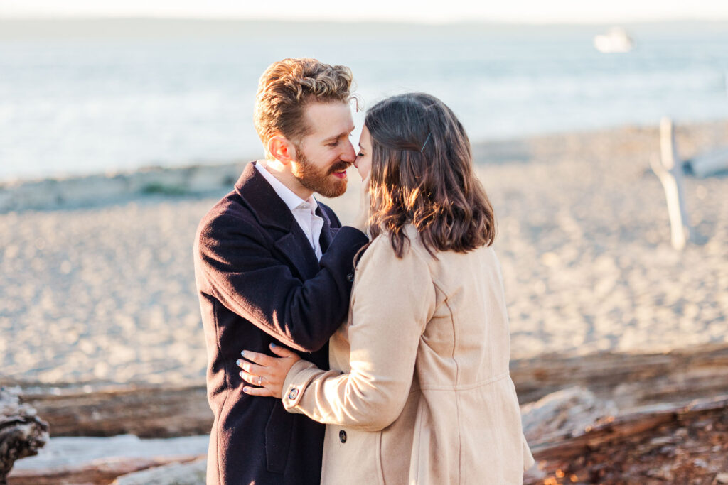 "Engaged couple sharing a romantic moment on a sandy beach with driftwood, the soft glow of sunlight highlighting their embrace as the calm ocean stretches into the distance.