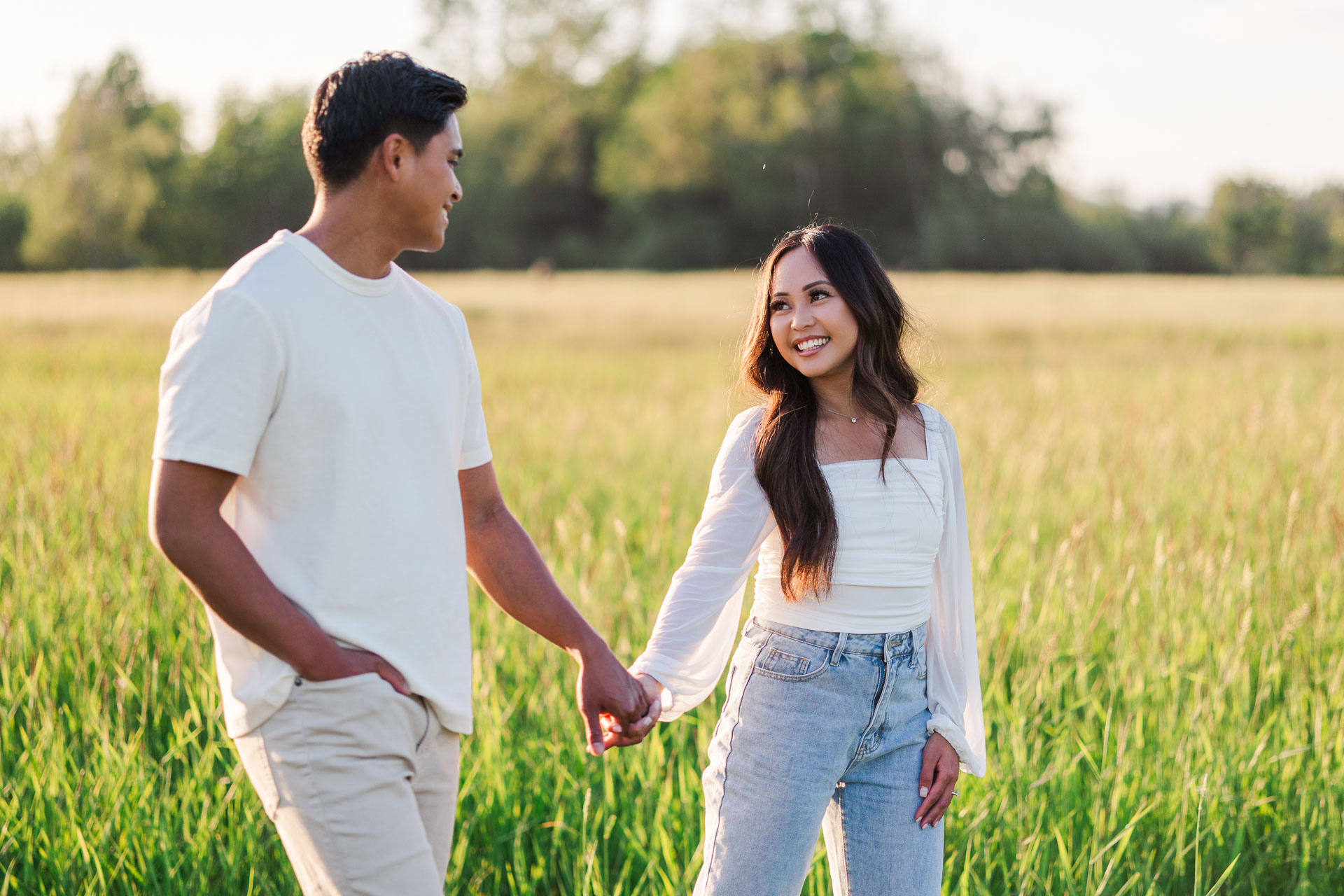 Engaged couple walking hand-in-hand through a sunlit meadow, sharing a warm smile and enjoying the golden hour glow amidst the tall grass in Elk Fields.