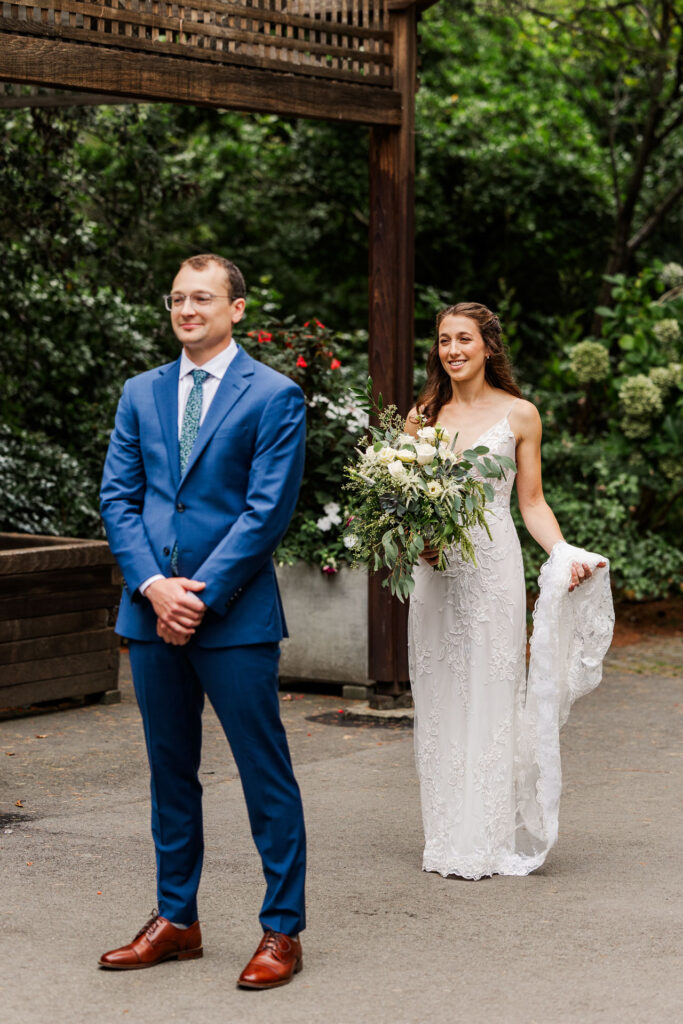 Bride approaching the groom for their first look, holding her bouquet and lace dress train, set against a garden backdrop with vibrant greenery.