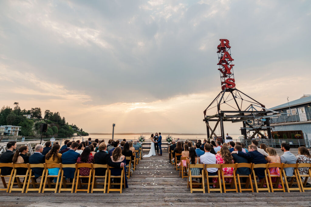 Couple exchanging vows during an outdoor waterfront wedding ceremony at Ray's Boathouse in Seattle, surrounded by guests seated on wooden chairs, with a stunning sunset sky and scenic water views in the background.