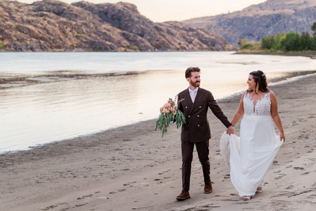 Bride and groom walking hand in hand along a serene lakeside beach with rugged cliffs and calm water in the background, showcasing a romantic and natural setting for wedding portraits.