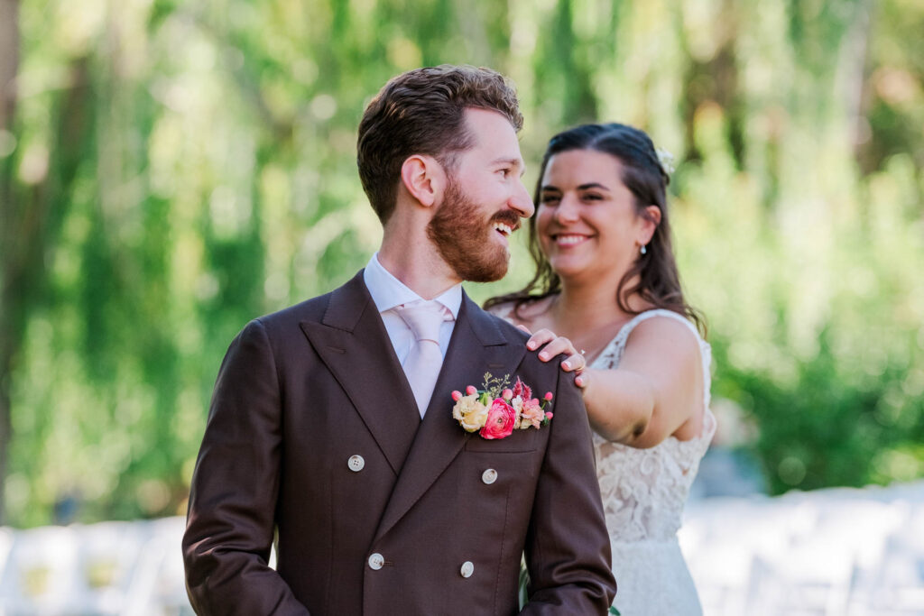 Bride smiling as she places her hand on the groom's shoulder during their first look, with the groom turning to smile back, surrounded by a lush green outdoor setting.