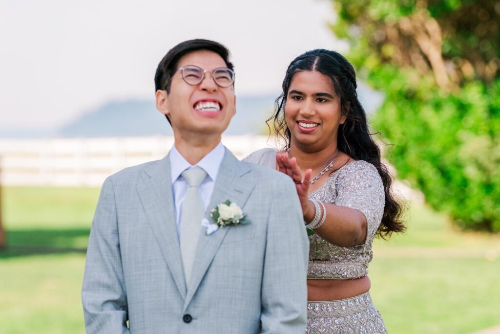 Groom grinning with anticipation as the bride approaches from behind during their first look moment, set outdoors with a bright, lush background.