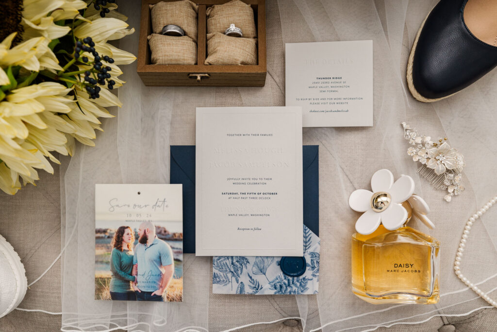 Flat lay of wedding details, including a wooden box with rings, wedding invitations, a save-the-date card, Daisy by Marc Jacobs perfume, pearl jewelry, and floral accents, styled on a soft neutral background.