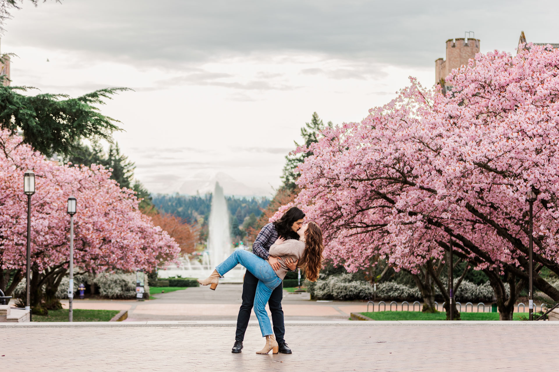 Couple sharing a playful dip and kiss beneath blossoming cherry trees at the University of Washington campus, with the iconic Drumheller Fountain and Mount Rainier visible in the background.